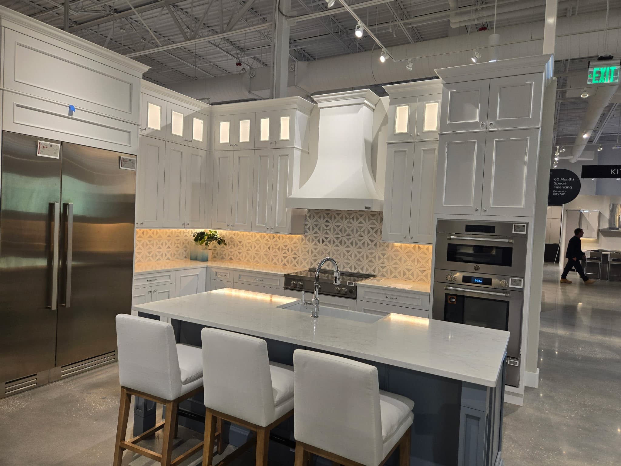 Modern white kitchen showroom featuring a marble island, stainless steel appliances, and under-cabinet lighting.