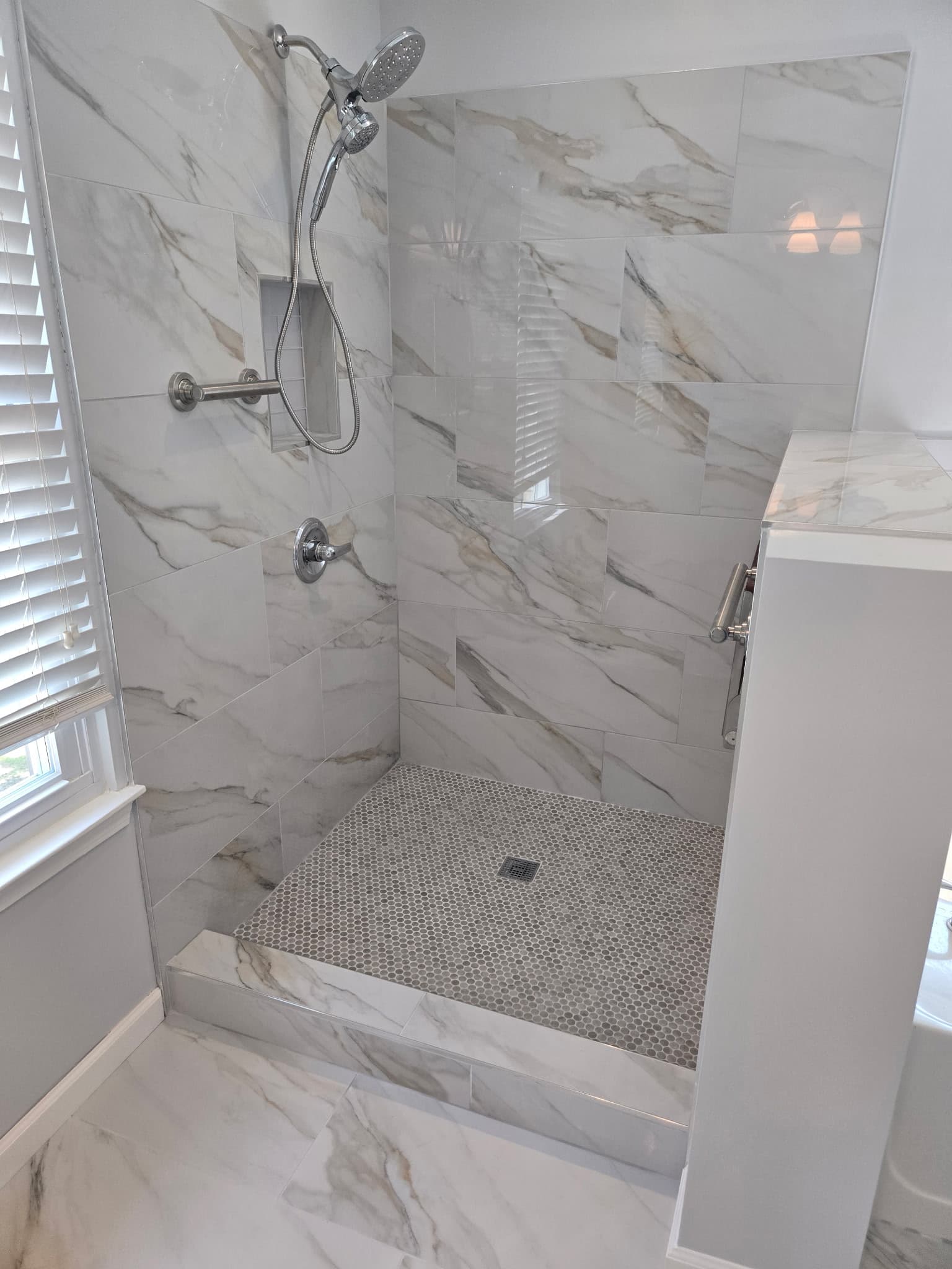 Modern walk-in shower with marble-patterned wall tiles, grey penny floor, and chrome fixtures.